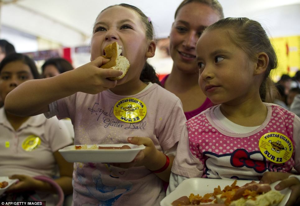 Two young people wearing pink. One person is eating a sandwich with great abandon; the other looks on in envy.