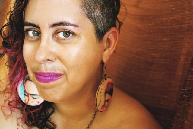 A woman with light brown skin, salt and pepper hair, and bright pink lips looks into the camera against a brown background. She is wearing large circular earrings featuring renderings of Frida Kahlo in either ear.