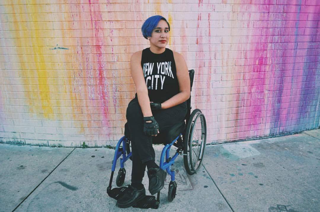 Annie, a young Latinx woman with blue hair in this photo, sits in her wheelchair with legs crossed. She is in front of a white wall that's been splashed with yellow, pink, and purple paint, and is wearing black head-to-toe, with a shirt that says New York City in white block letters.