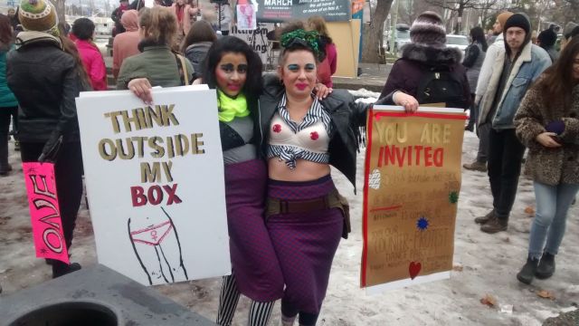 Two people pose for a photo while holding protest signs; one reads "think outside my box" and the other is an invite to a counter-protest dance party