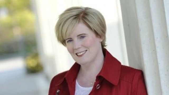 A white woman with light blonde hair in a red jacket and white shirt leans against a white pillar.