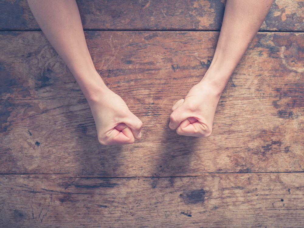 A white woman's clenched fists on a wooden table, shot from above.