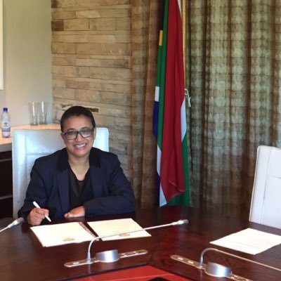 A woman with closely shaved black hair and black rimmed glasses sits behind a desk in a government office smiling at the camera. There is a South African flag in the background and various papers are on the desk. She is wearing a navy blue blazer and black shirt.