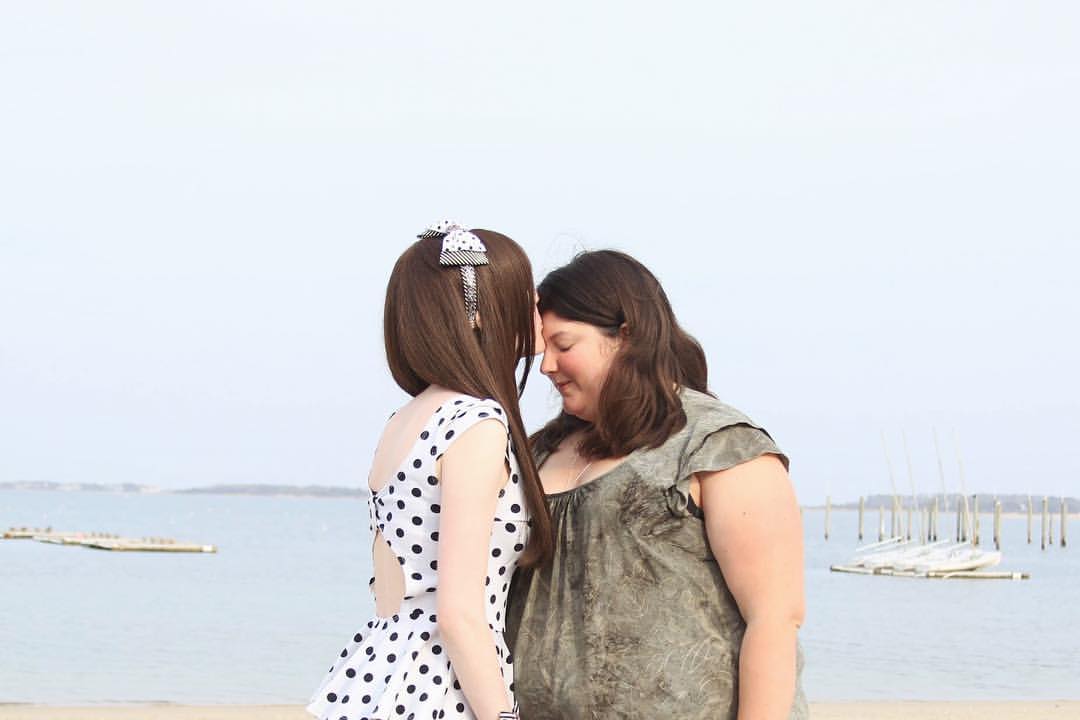 Two people stand on the shore against the background of the sea. They both have brown hair and are wearing dresses; the person on the left is in a white dress, and the person on the right is in green. The person on the left is leaning in to kiss the other one's forehead.