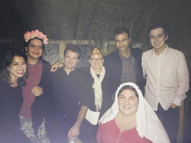 A group of seven people, six standing in a back row and one crouching down in front, smile into the camera. Some of them are wearing parts of Halloween costumes, including a veil, a gold crown, and a crown of flowers.