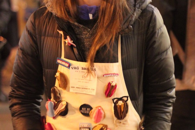 A person wearing a pink "pussy hat" and an apron with lots of buttons and sculpted vulvas pinned to it