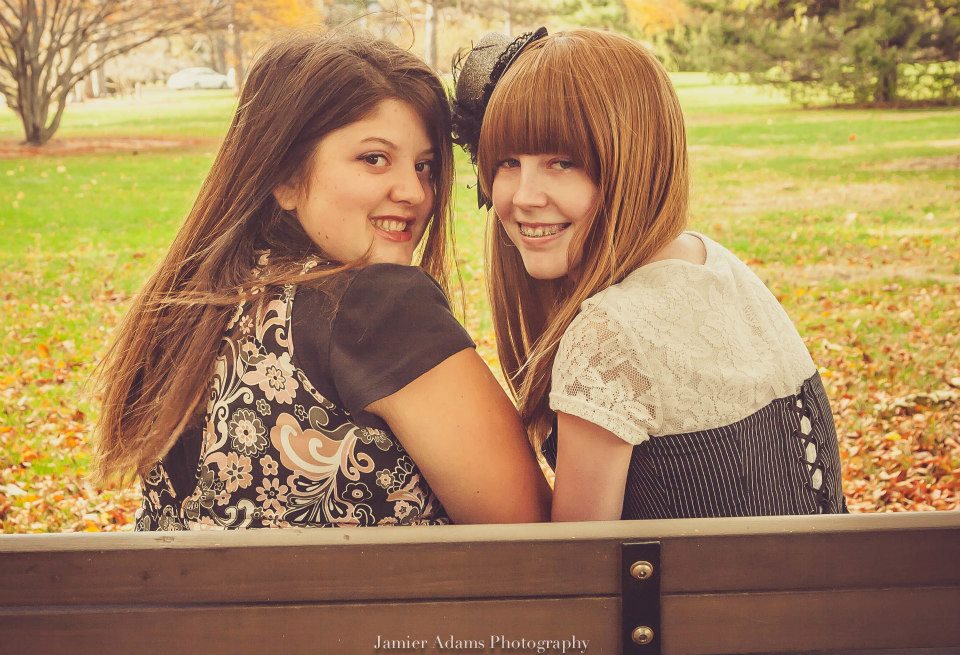 Two people sit on a bench smiling into the camera by looking back over their shoulders. The person on the left is in a black shirt with a patterned top, and the one on the right is in a white lacy top with a black corset over it. They both have brown hair, though the one on the left has darker hair than the one on the right.