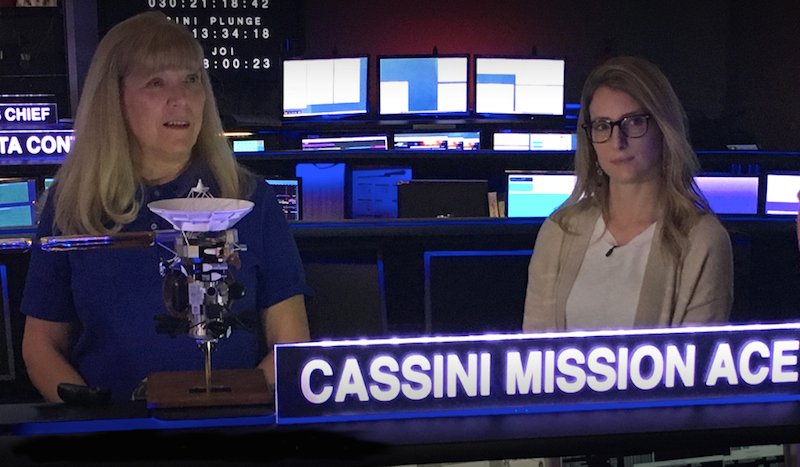 Two white women stand next to each other in a mission control room, with the text "Cassini Mission Ace" lit up in front of them. Linda Spilker is blonde and wears a blue polo shirt; Molly Bittner has glasses and a tan cardigan. There are numerous monitors lit up behind them.