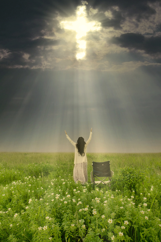 Rear view of a woman in a long white skirt and top standing in a meadow next to an empty manual wheelchair, with her arms raised toward a dark sky. A cross can be seen through the clouds and is bathing her in a beam of light.