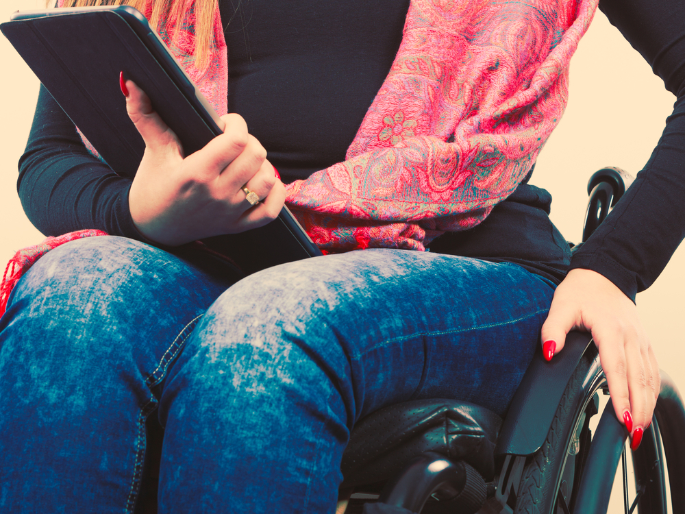 A woman sits in a manual wheelchair holding a tablet computer. She is wearing blue jeans and a pink scarf and has long red nails. The photo only shows her from the neck down, no head.