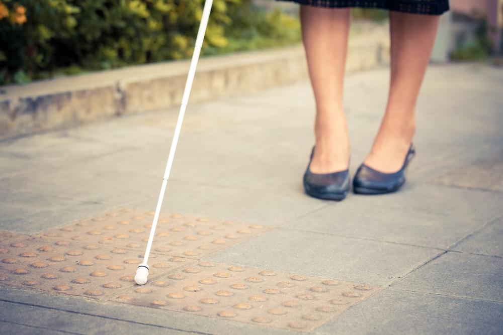 A woman's feet walking along a sidewalk. She has a white cane out in front of her. You can only see her black shoes, part of her calves, and the tip of the cane.