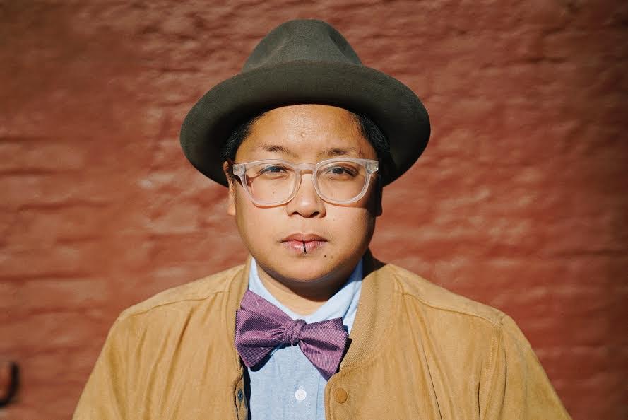 A Filipinx person looks into the camera against a brick wall background. They are wearing clear framed glasses, a grey bowler hat, a mustard yellow jacket and blue shirt, and a purple bow tie.