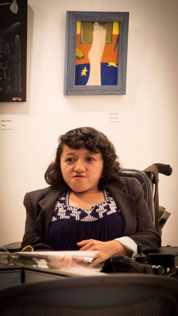 A young Asian woman with shoulder-length hair and wearing a black shirt with white trim sits in her wheelchair looking at the camera.