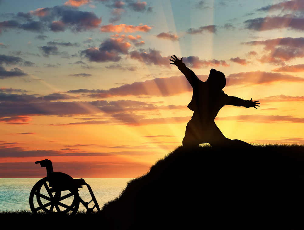 In silhouette against a sunset background, a person drops to their knees on a small hilltop and raises their arms out wide. Their manual wheelchair sits empty at the bottom of the hill.