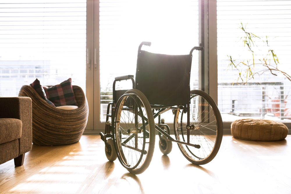 A black manual wheelchair sits facing a large window, with light streaming in over the chair from outside.