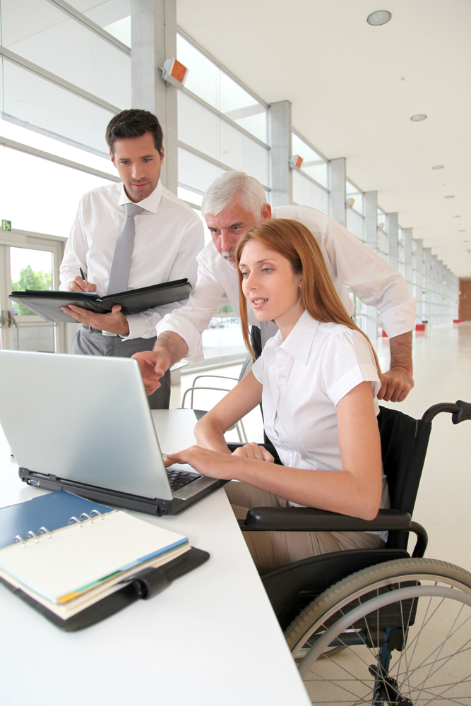 A woman with reddish brown hair in a white short-sleeved blouse looks at a laptop computer screen while two men, one older and one younger, stand behind her. The older man is pointing at something on the screen while the younger one is taking notes. The woman is sitting in a manual wheelchair.