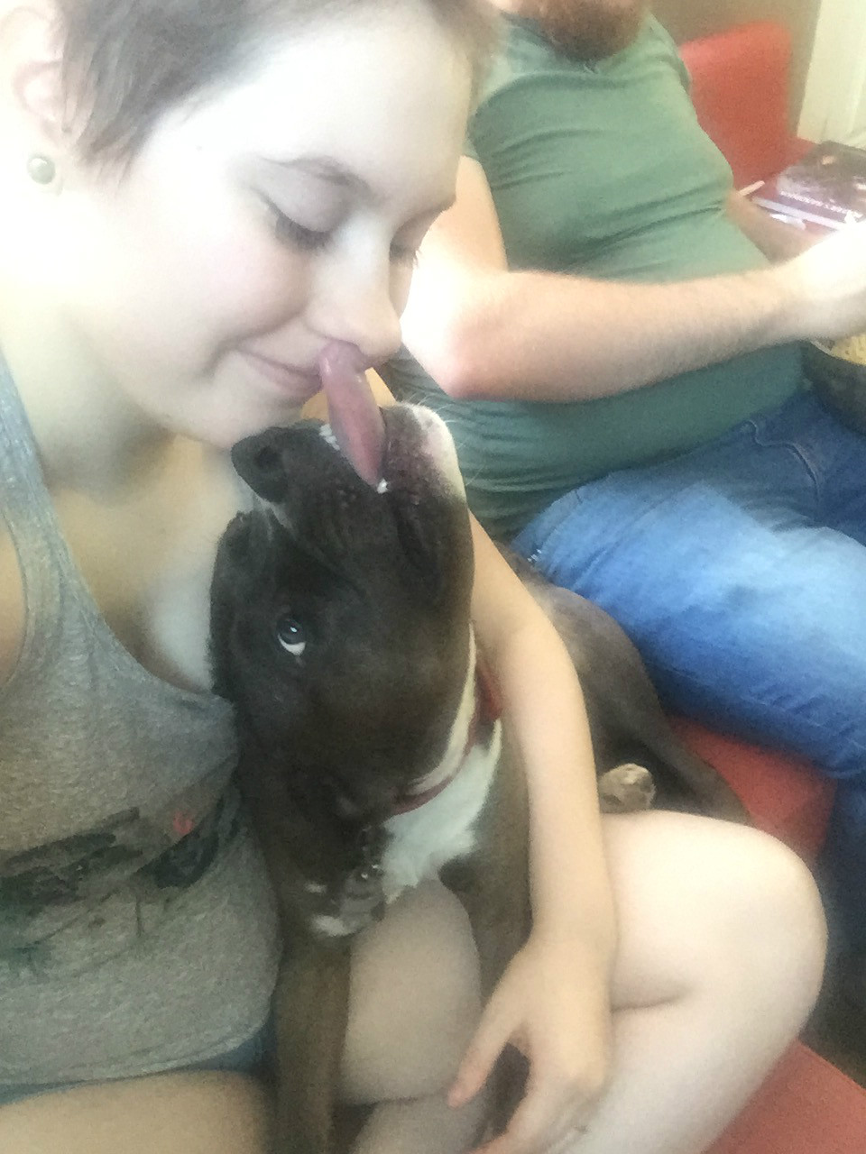 A pale-skinned person in a gray tank top smiles as a black and white dog sits in their lap and licks their face.