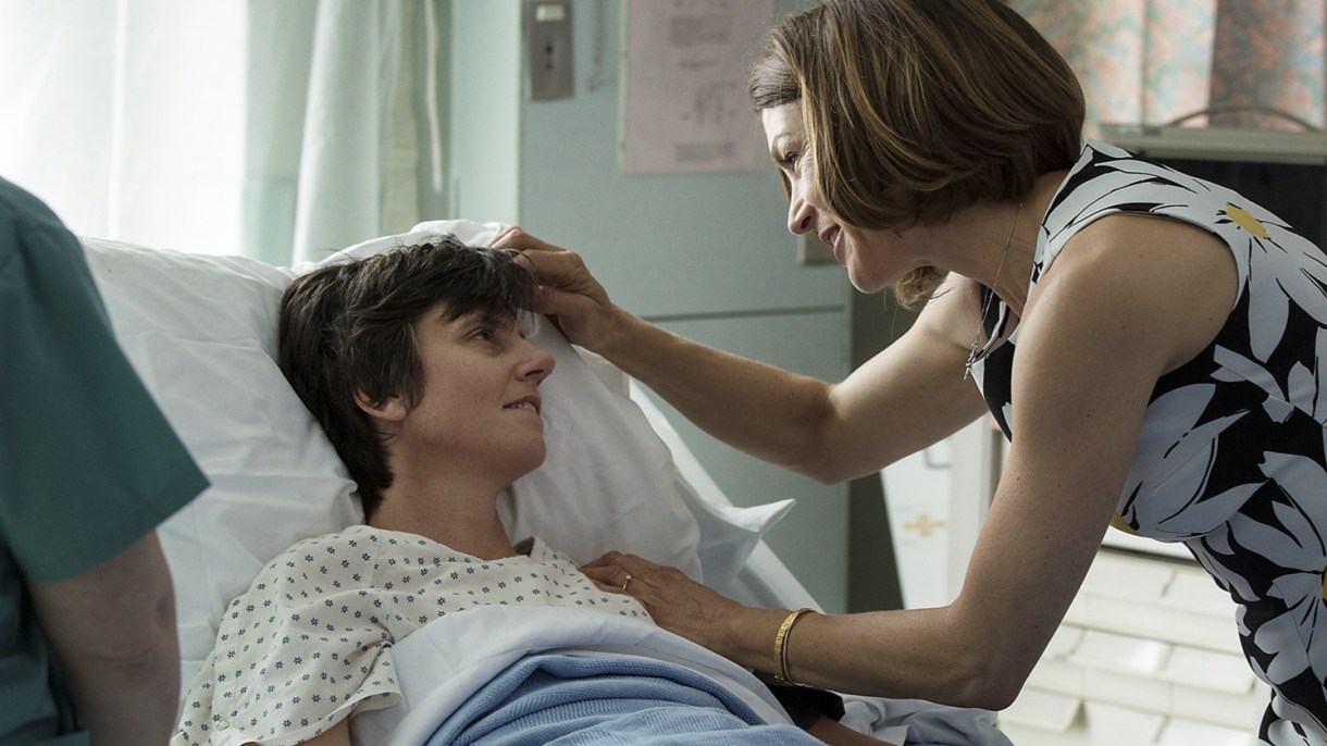 Tig, played by Tig Notaro, is in a hospital bed. A woman in a dress is touching her forehead lovingly.