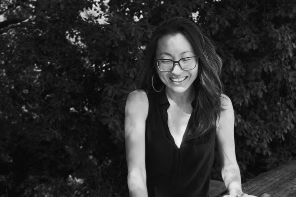 An Asian woman with long dark hair and square glasses smiles down toward the ground. The photo is in black and white, and she is wearing a sleeveless black top with a v-neck.