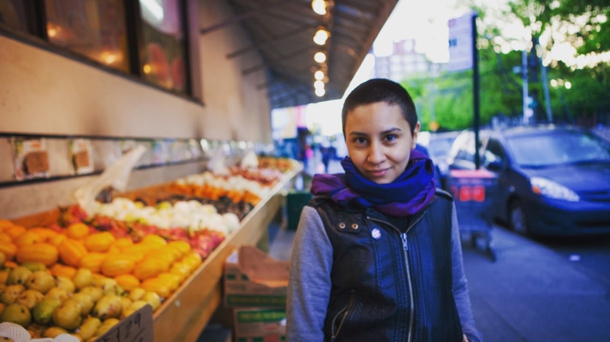 A person with a closely shaved head wearing a grey shirt, purple and blue infinity scarf, and black leather vest stands next to a sidewalk produce stand.