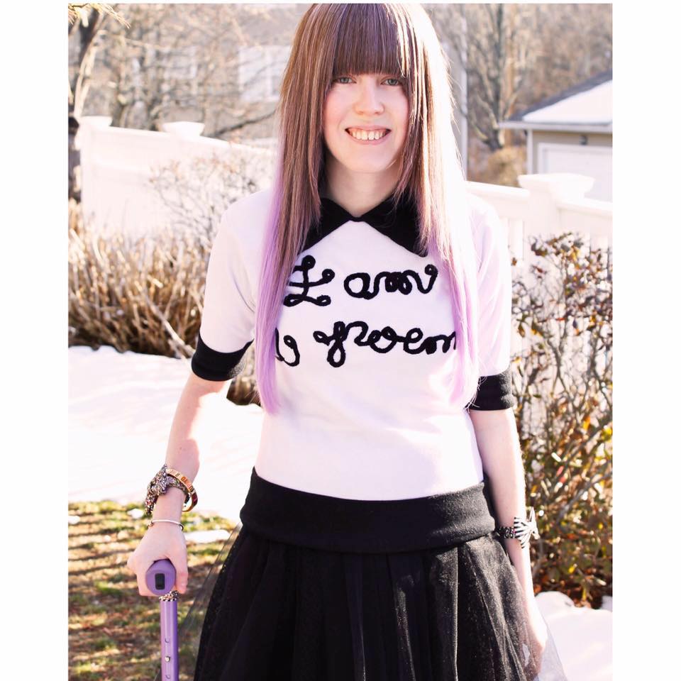 A person with long brown hair dyed purple at the tips holds a sparkly purple cane in their right hand.