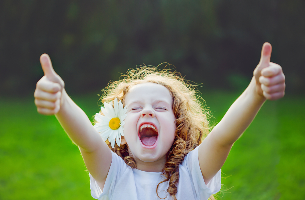 A young girl with pale skin and red hair wearing a white shirt and a flower behind her ear gives an enthusiastic thumbs up with both hands.