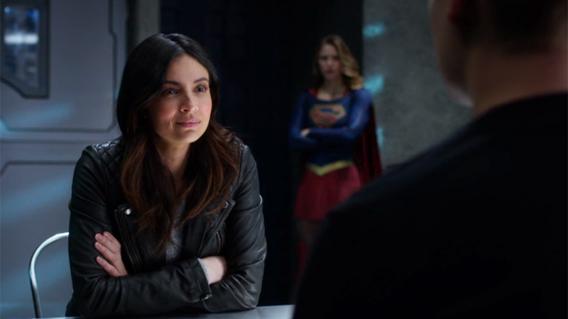 Maggie sits at the table, Supergirl stands watch behind her