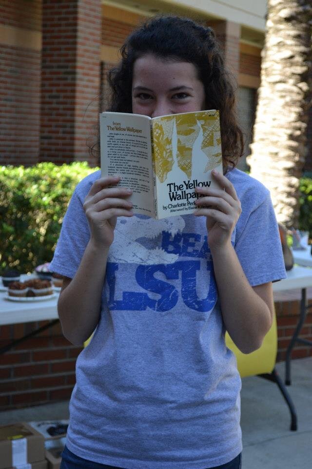 A young white woman with curly brown hair smiles from behind the book The Yellow Wallpaper.