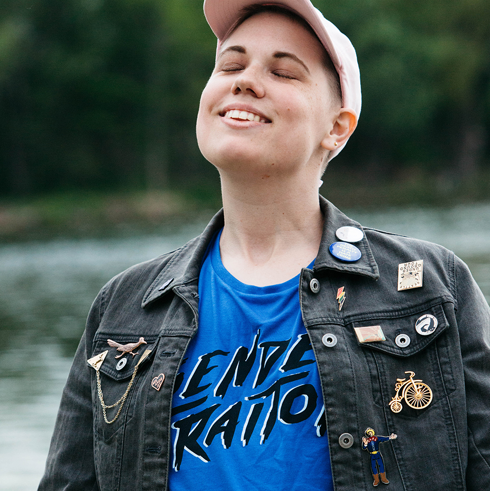 Audrey basking up the sun in their Gender Traitor Tank, black denim jacket, with so many enamel pins