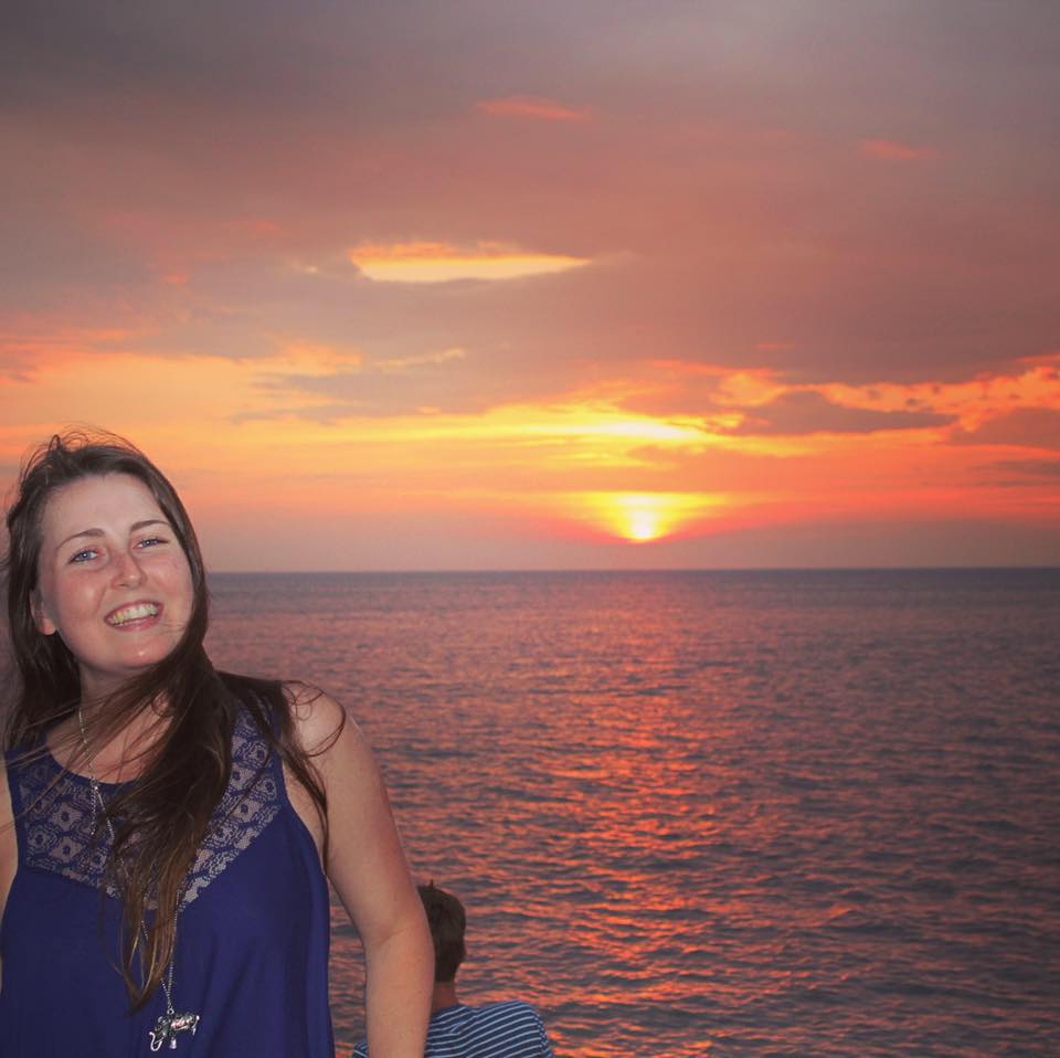 A young white woman with long brown hair smiles in front of a sunset over the ocean.