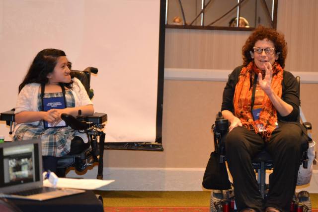 Sandy sits next to the author Simi Linton on stage at a conference. Sandy is on the left in a blue dress and white sweater, while Simi is on the right in all black except for an orange scarf around her neck.