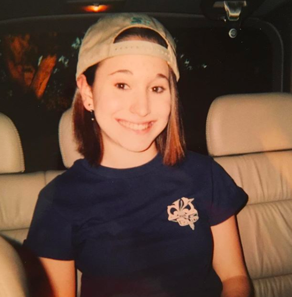 A photo of Gaby Dunn in a backward baseball cap and a blue t-shirt, possibly in a car or van? Definitely smiling!