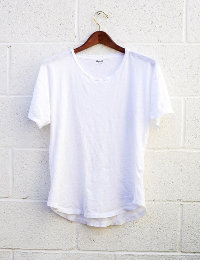 A white short-sleeved t-shirt, hanging against a white brick wall.