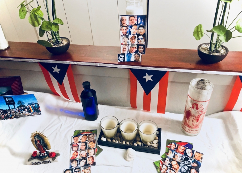 A home photograph of an altar built in a living room with a collection of white table cloth, candles, and Puerto Rican flags. 