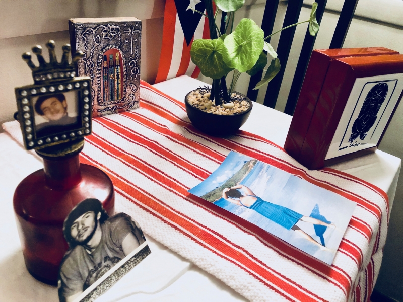A home photograph of an altar built in a living room with a collection of white table cloth, candles, and Puerto Rican flags. 