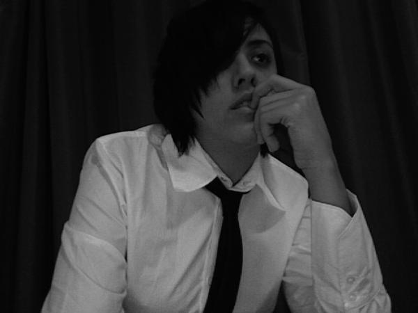 A picture of your host, Carly Usdin, from 2007. They are wearing a white button-up shirt and a skinny black tie. They have an extremely gay asymmetrical haircut and have one finger demurely in their mouth as they stare off into the distance.