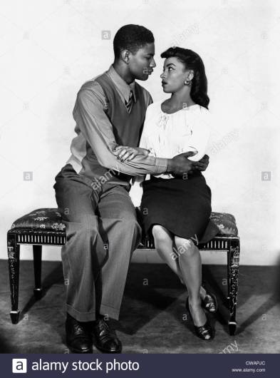 A black-and-white photo, featuring a Black man (wearing trousers, a button-up and tie, and a vest) locking arms with a Black woman (wearing a black skirt and white top) as they sit on a bench and stare into each others' eyes. This is a stock photo that appeared in the background of this episode of The L Word.