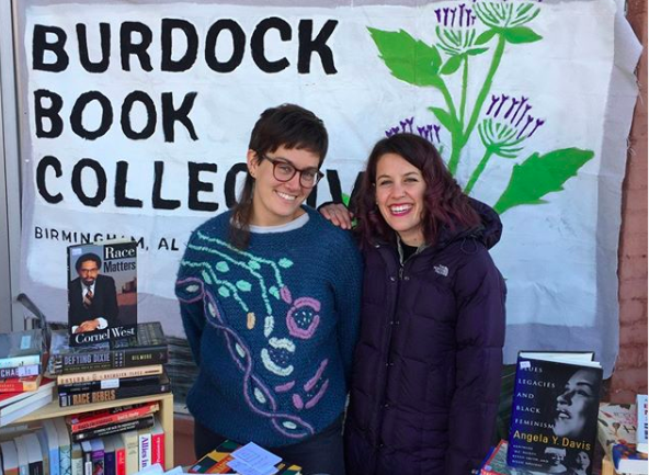 Two people stand in front of a sign reading Burdock Book Collective, smiling at the camera