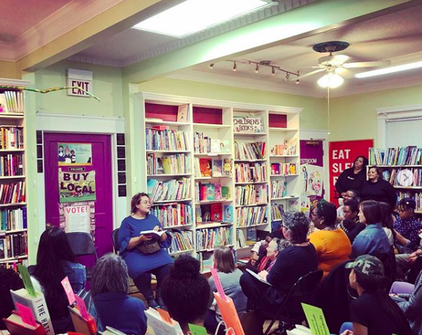 A seated person reads from a book to an assembled gathering of listeners, with bookshelves and a sign that reads BUY LOCAL on the walls behind her