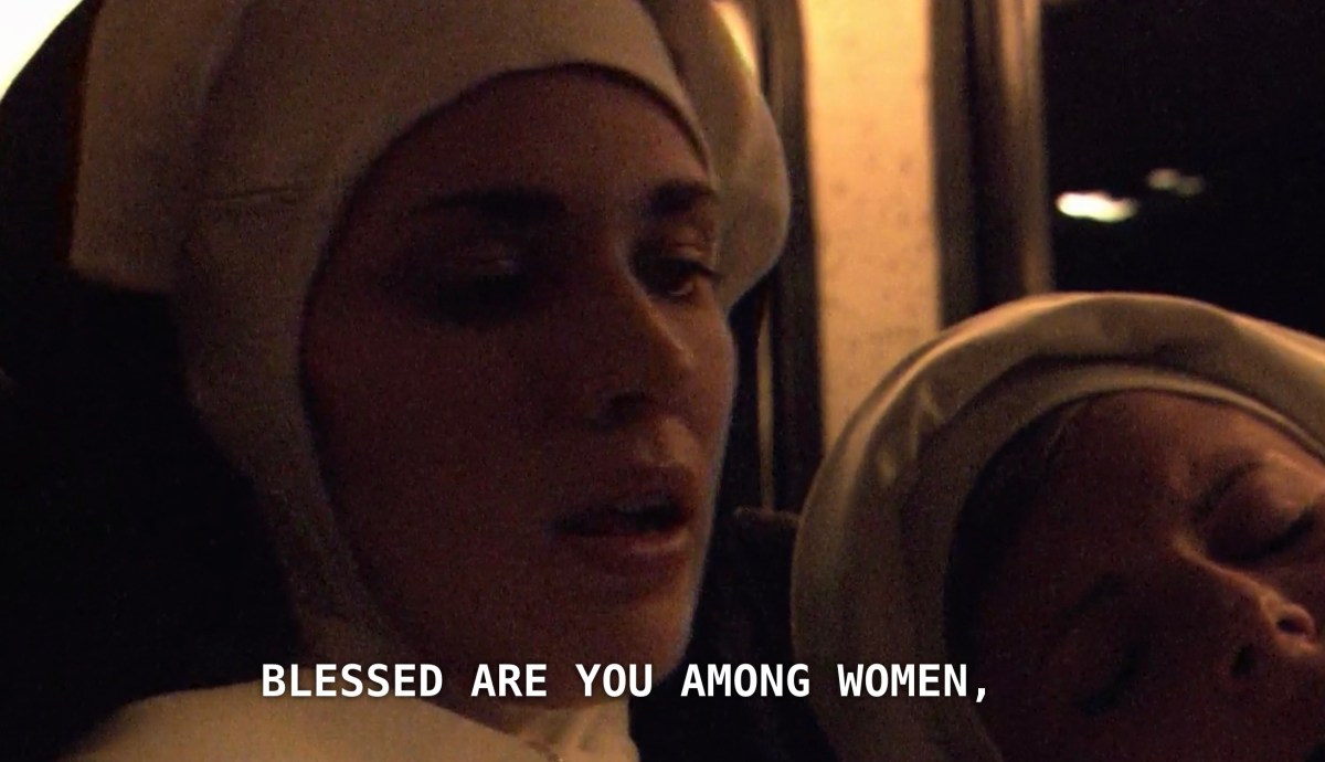 A nun riding on a charter bus, wearing nun attire and her mouth slightly open, saying "blessed are you among women." Another nun sits asleep next to her.