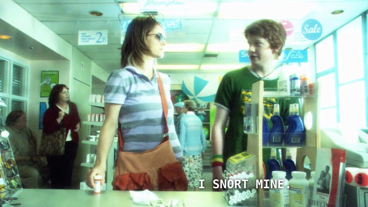 Alice (wearing a very 2000s purple and gray striped polo shirt and enormous brown corduroy bag) stands at the counter of a pharmacy. She is anxiously awaiting the pharmacist to retrieve her new meds. A young red-head boy (wearing a green and yellow t-shirt) says to her, "I snort mine."