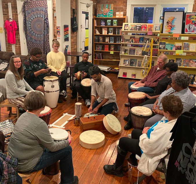 A group of a dozen or so people sit in a circle with drums before them, a row of bookshelves behind them