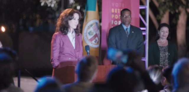 Bette in a mauve pantsuit stands at a podium speaking to a crowd at a mayorial race event.