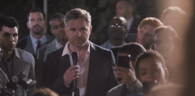 A man in a white shirt and blazer speaks on a microphone, surrounded by people