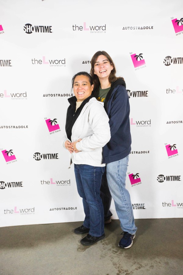 Two people posing "prom style" in front of a promo poster for "The L Word: Generation Q." The person standing behind is wearing jeans and a blue hoodie. That person has their arms around a person standing in front of them, who is wearing blue jeans and a white jacket.