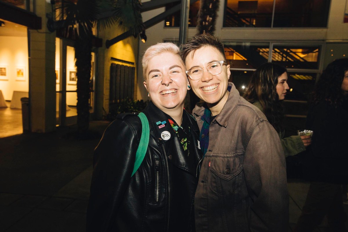 Two people pose for a picture, standing so close their cheeks are touching. The one on the left has short bleach blonde hair and a yellow jacket. The person on the right is wearing a brown jacket and glasses, and has short brown hair.