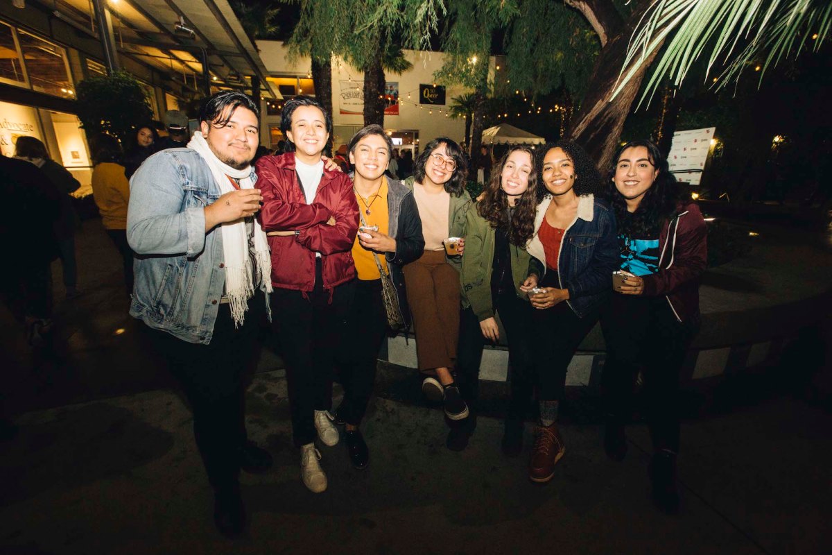 A group of 7 people stand in a line posing for a picture. Some are holding cocktail cups of beverages. 