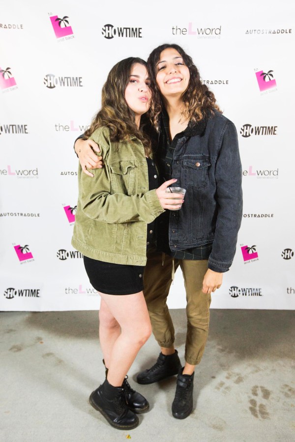 Two people with their arms around each other in front of a promo poster for "The L Word: Generation Q." The person on the left is wearing a black mini skirt and green jacket. The person on the right has tan pants and a black denim jacket.