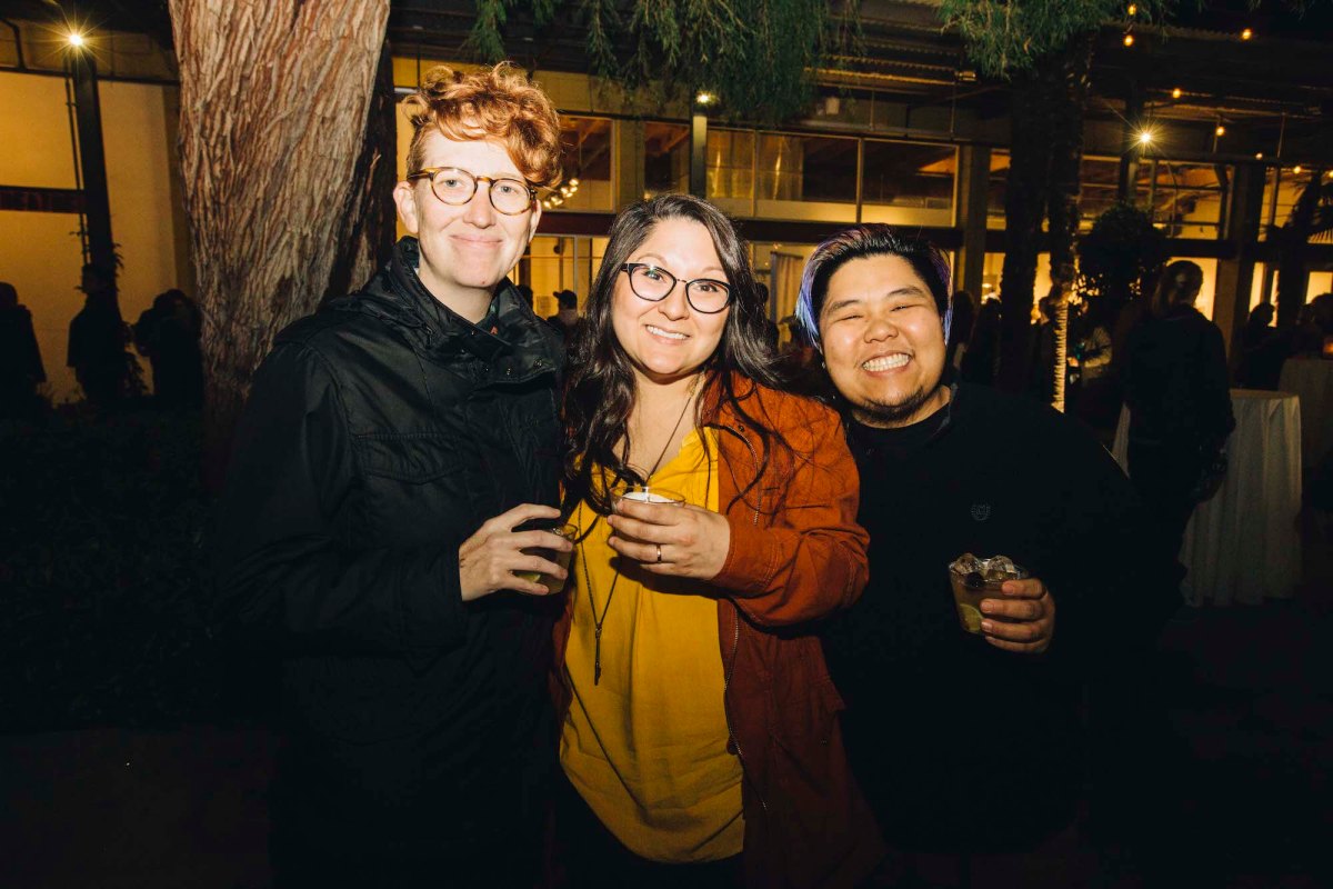 Three people posing for a photo. The one on the left has short red hair, glasses, and a black shirt. The one in the middle is wearing a yellow shirt and a red jacket, glasses, and long brown hair. The one on the right is wearing a black shirt, has short dark hair, and facial hair.