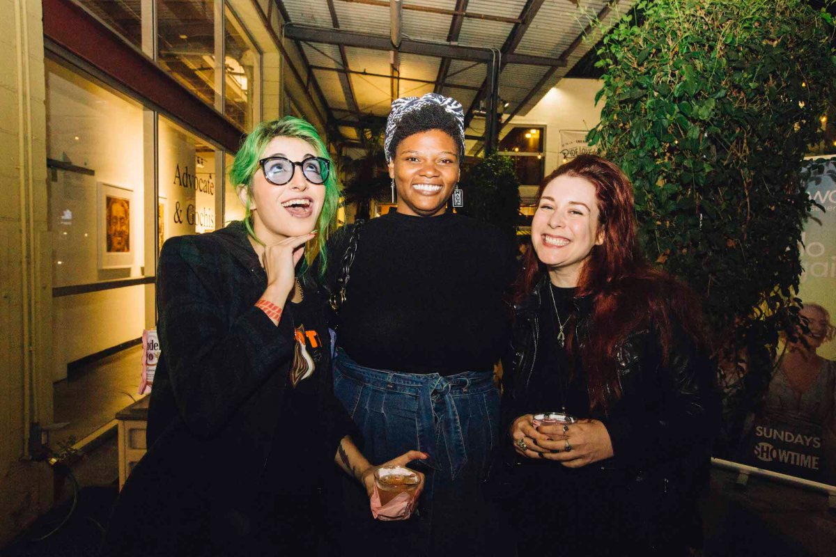 Three people posing for a photo. The one on the left has green hair and is wearing all black and glasses. The person in the middle is wearing a denim skirt, black top, and a headscarf. The person on the right has long dark red hair and is wearing all black. 